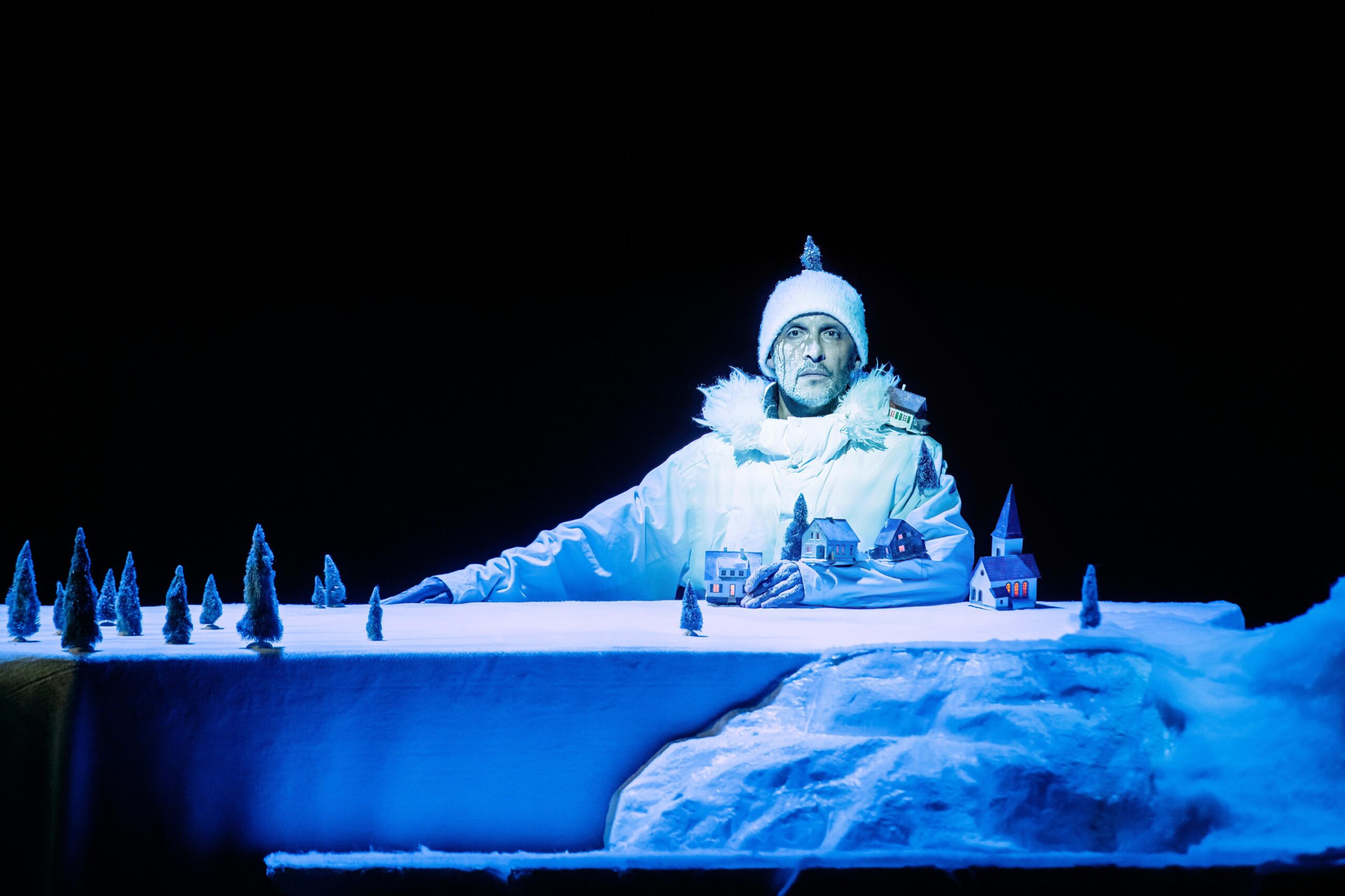 Un homme en veste de ski blanche regarde l'objectif. Il porte un bonnet blanc et sur son visage, il y a de la neige. Il est assis à une table recouverte d'un drap blanc et de petits sapins enneigés. Son bras repose sur la table, il y a de petites maisons collés sur sa manche de veste, qui font comme un village d'hiver.