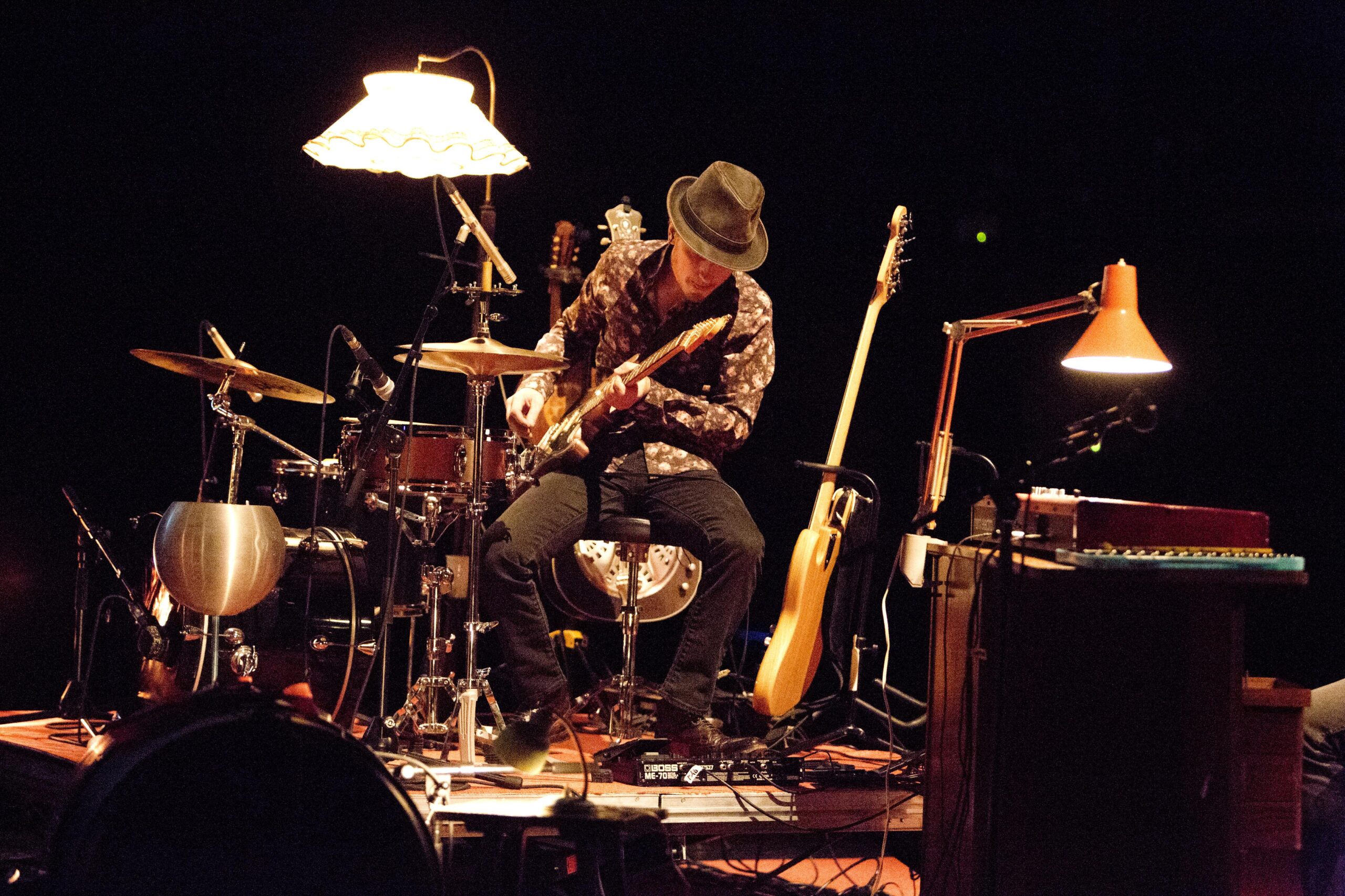 Un homme avec un chapeau est assis au milieu de plein d'instruments. Il tient une guitare dans les mains.