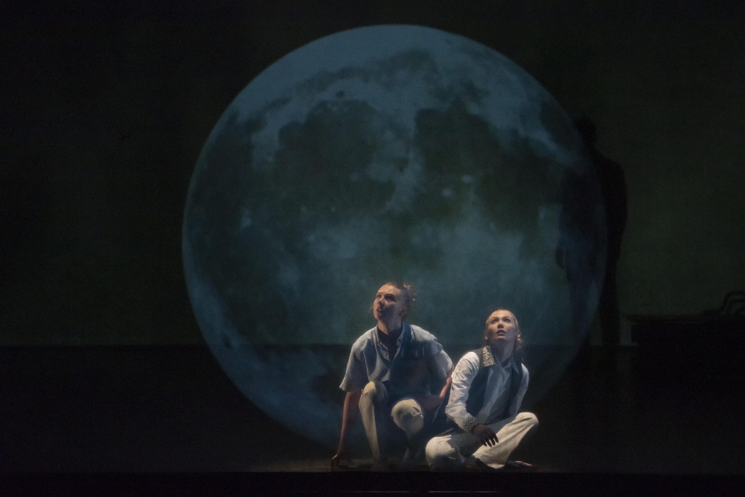 Deux personnes sont assises au sol et regardent vers le ciel. Une projection donne l'illusion qu'elle sont dans une bulle, qui ressemble à la Lune.