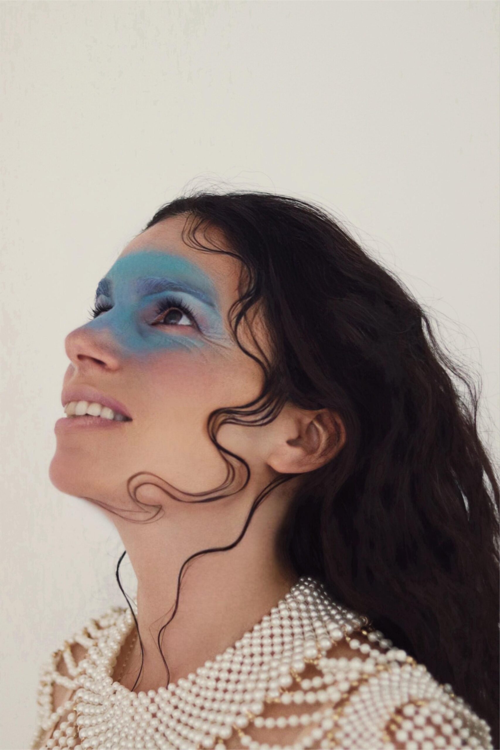 Yael Naim regarde vers le ciel. Elle est vêtue d'un haut en perles blanches. Ses cheveux tombent vers l'arrière. Sur son visage, autour de ses yeux, elle porte comme un masque de maquillage, bleu océan.