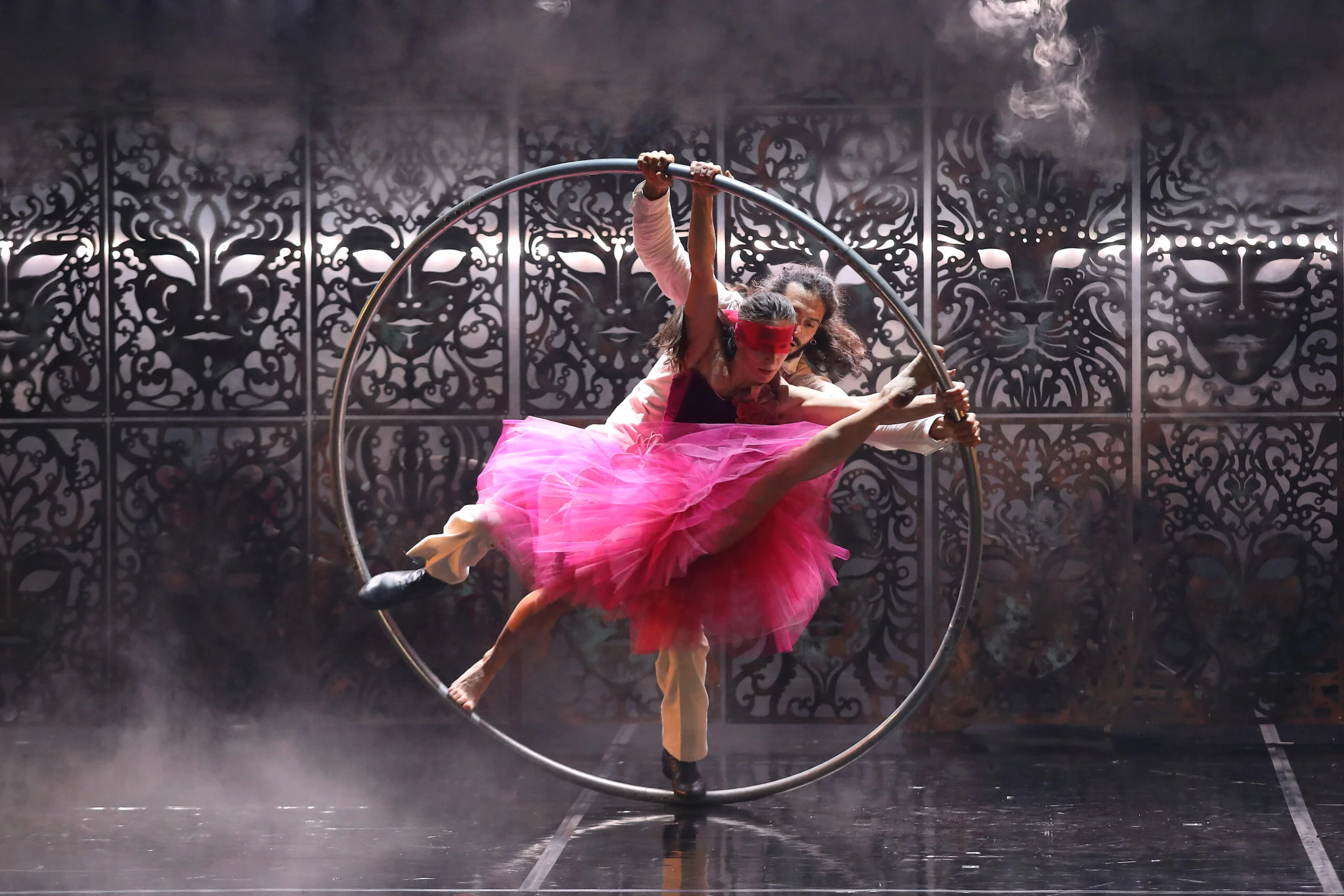 Une femme et un homme tiennent en équilibre dans un cerceau en mouvement. La femme fait une sorte de grand écart. Elle porte un grand tutu rose vif et un bandeau de la même couleur sur ses yeux. L'homme porte un complet beige.
